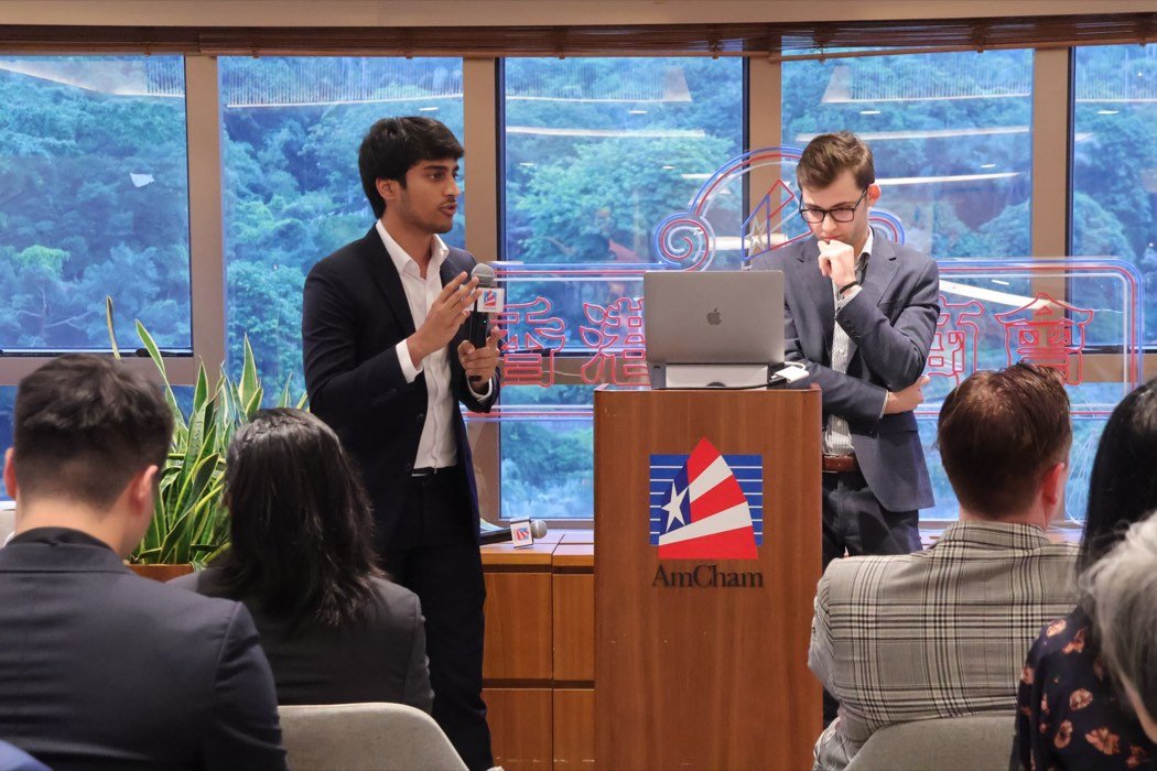 A presenter answers a question into a microphone beside an AmCham lectern, with a colleague and a seated audience.